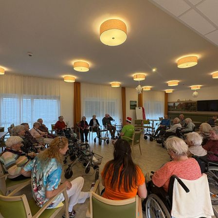 Ältere Menschen sind in einem Raum mit Rollstühlen und Gehhilfen versammelt. Es gibt mehrere Lampen an der Decke. Einige tragen Mützen, und einer trägt einen Weihnachtsmannhut.