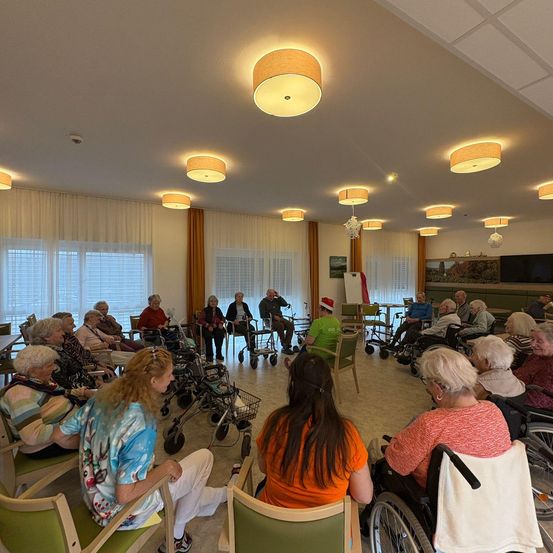 Ältere Menschen sind in einem Raum mit Rollstühlen und Gehhilfen versammelt. Es gibt mehrere Lampen an der Decke. Einige tragen Mützen, und einer trägt einen Weihnachtsmannhut.
