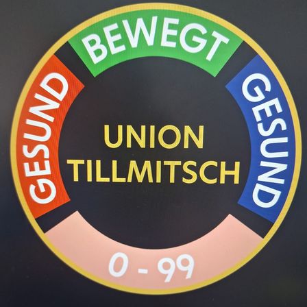 Ein rundes Schild zeigt den Text 'UNION TILLMITSCH' mit den Zahlen '0-99' in der Mitte, umgeben von mehrfarbigen Abschnitten, die mit 'GESUND' beschriftet sind.