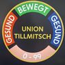 Ein rundes Schild zeigt den Text 'UNION TILLMITSCH' mit den Zahlen '0-99' in der Mitte, umgeben von mehrfarbigen Abschnitten, die mit 'GESUND' beschriftet sind.