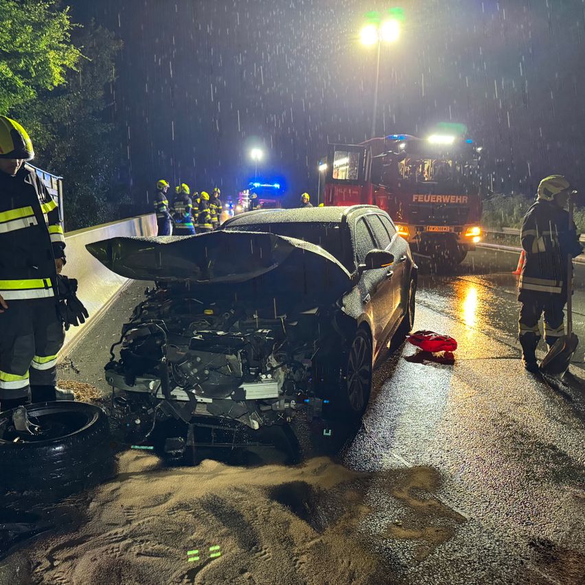 Ein Auto ist auf einer regnerischen Straße in der Nacht beschädigt. Feuerwehrleute in gelben Uniformen sind um das Auto herum. Ein Feuerwehrwagen ist hinter dem Auto geparkt.