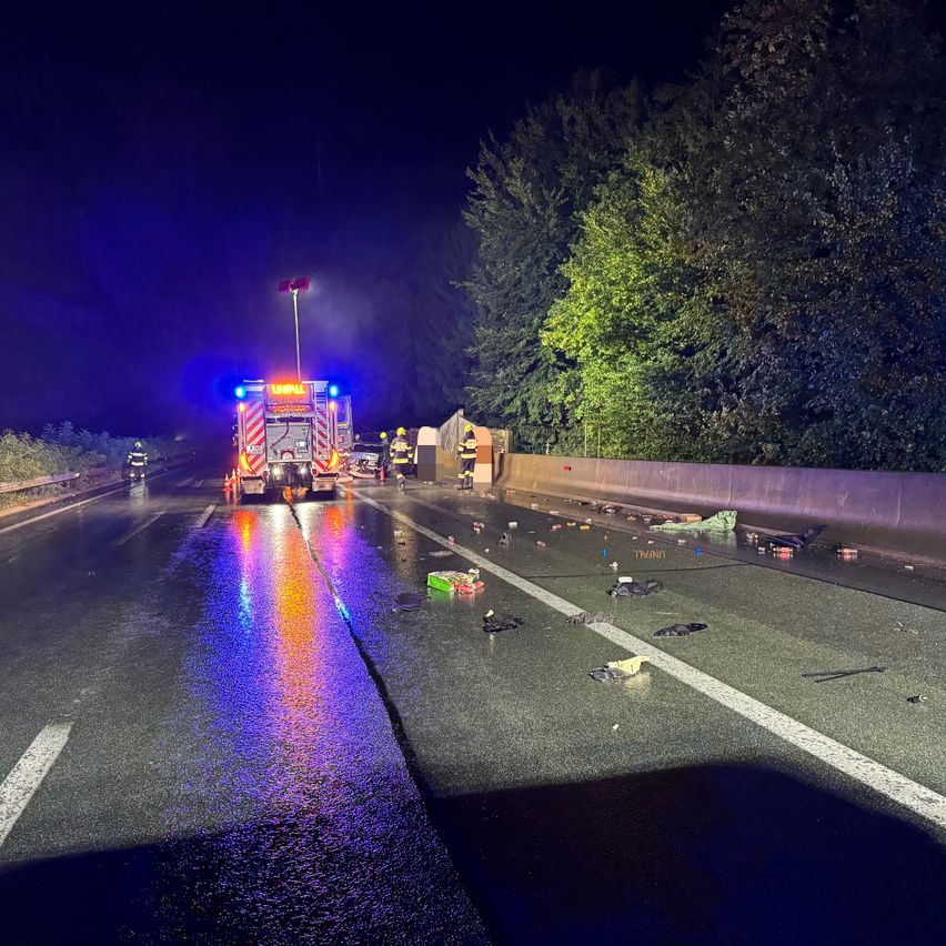 Eine Autobahnszene bei Nacht mit Einsatzfahrzeugen und Trümmern auf der Straße. Die Straße ist nass, und Notleuchten blinken. Bäume und eine Barriere sind sichtbar.