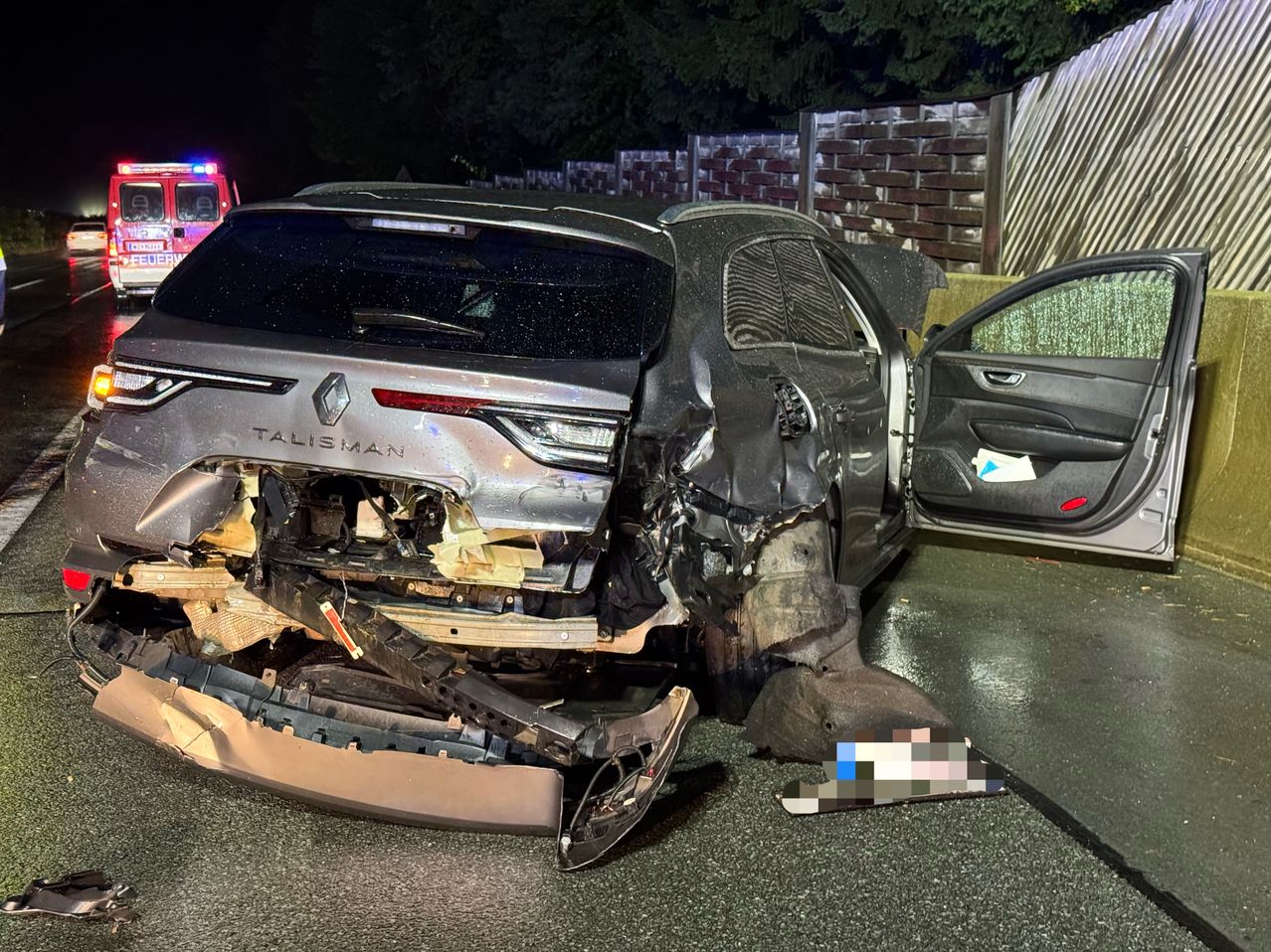 Ein schwer beschädigter Renault Talisman ist bei Nacht zu sehen, mit zerquetschter Front, eingeschalteten Lichtern und einem Feuerwehrwagen im Hintergrund.