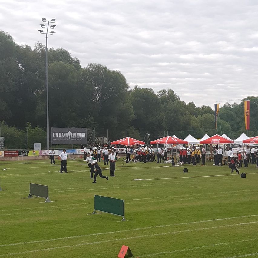 Eine Gruppe von Menschen befindet sich auf einem Fußballfeld, die an einem Trainingslager teilzunehmen scheint. Sie tragen weiße Uniformen. Einige von ihnen halten einen Fußball. Zelte stehen am Rande des Feldes.