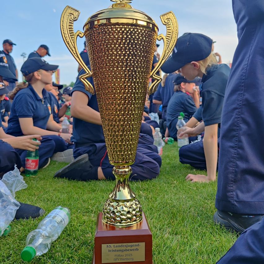 Ein goldener Pokal steht auf einem Rasenfeld, mit Leuten in blauen Uniformen, die sitzen und stehen. Eine Frau schaut auf den Pokal. Wasserflaschen und eine Plastiktüte sind in der Nähe.