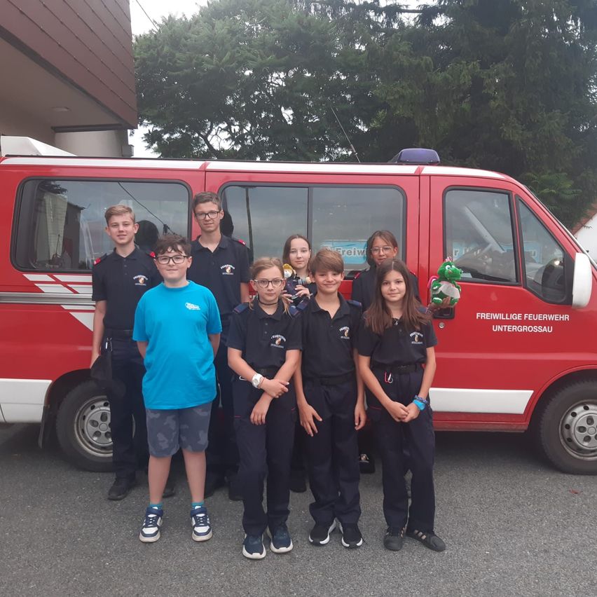 Eine Gruppe von Kindern in Uniformen posiert vor einem roten Feuerwehrauto. Im Hintergrund sind Bäume.
