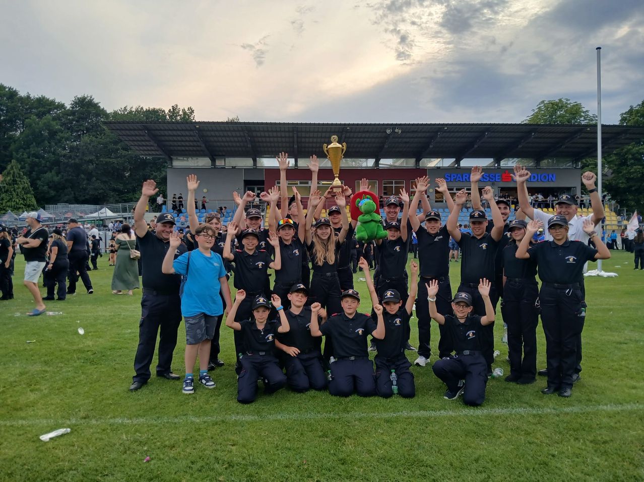 Eine Gruppe von Menschen posiert auf einem Feld für ein Foto, während sie einen Pokal hält. Sie tragen schwarze Uniformen, einige mit Brillen. Im Hintergrund ist ein Stadion mit einer rot-weißen Struktur zu sehen.