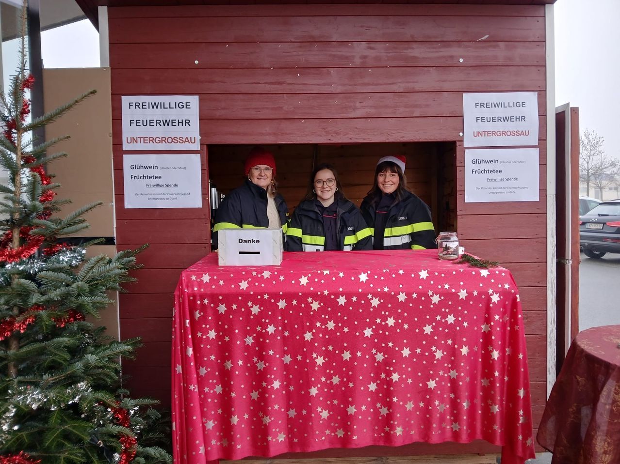 Drei Frauen in Feuerwehruniformen stehen hinter einem Schreibtisch in einer Feuerwache und lächeln für ein Foto. Vor ihnen stehen ein Weihnachtsbaum und eine Box mit der Aufschrift 'Danke'.