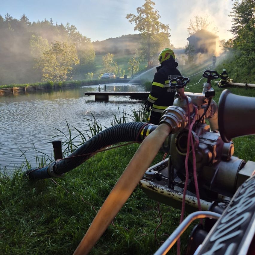 A firefighter is spraying water into a river from a boat. Trees and a house are in the background.