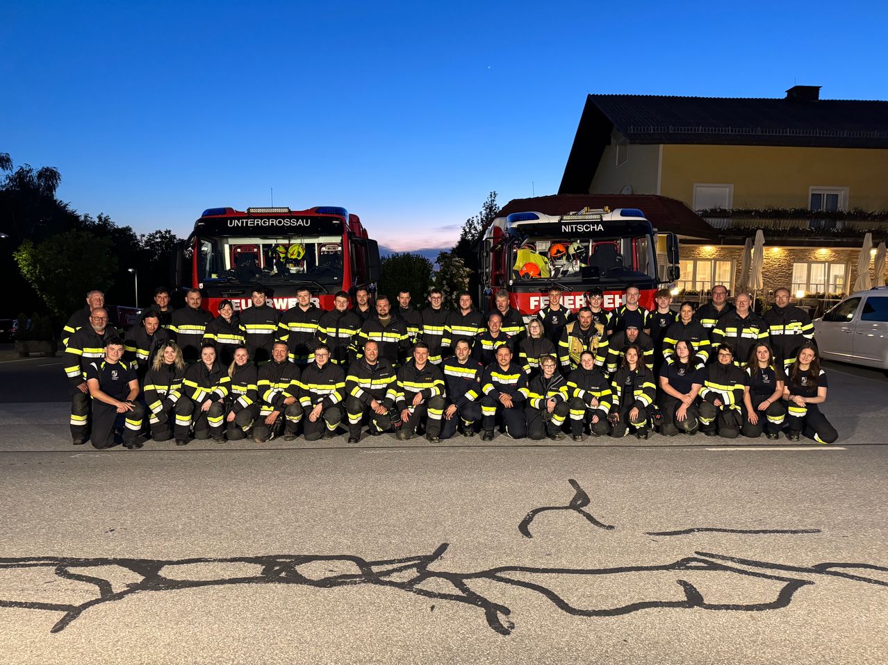 A group of firefighters is posing for a photo with two fire trucks in the background. The word Untergrossau is on one of the trucks.
