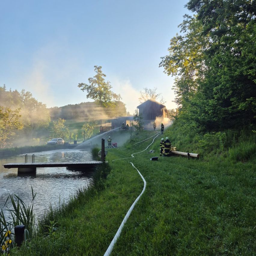 Firefighters respond to a house fire with smoke visible. They use a hose from a lake nearby. Trees and a bridge are in the background.