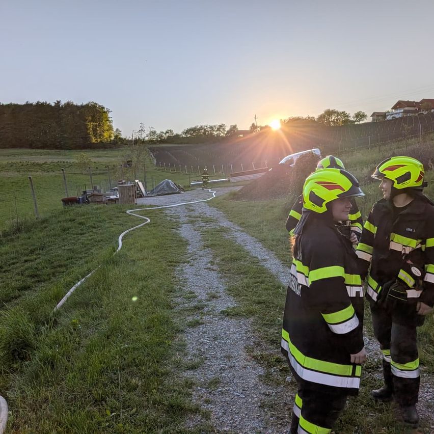 Three firefighters in yellow and black uniforms are standing on a gravel path. The sun is setting in the background.