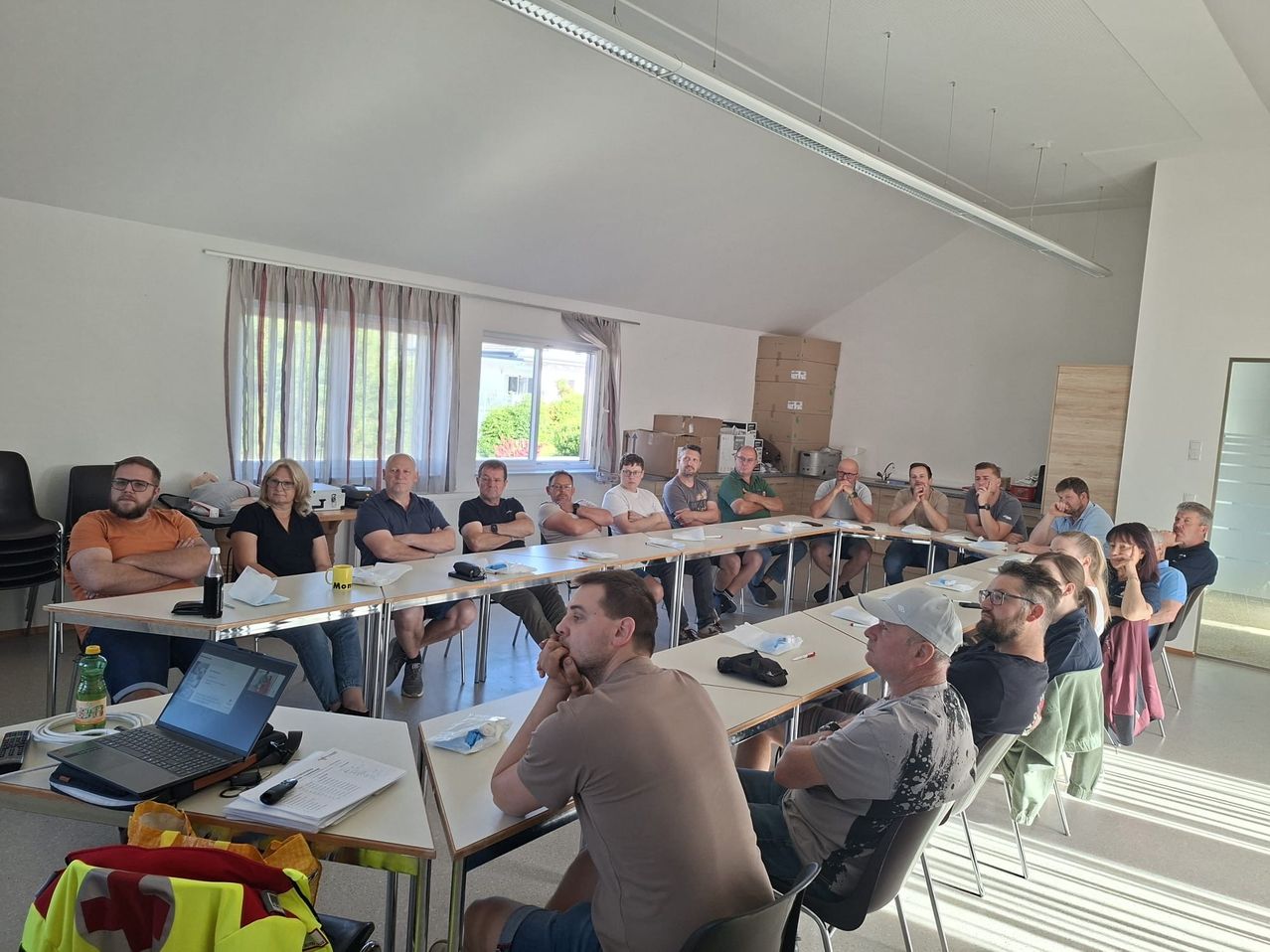 Eine Gruppe von Menschen sitzt in Stühlen um einen Tisch in einem Besprechungsraum, einige mit Brille. Ein Laptop steht auf dem Tisch, und eine Handtasche ist auf dem Stuhl. Ein Mann trägt eine Uhr, und eine Frau trägt eine Armbanduhr. Eine Tafel ist an der Wand, und einige Lagerboxen sind auf dem Regal platziert.
