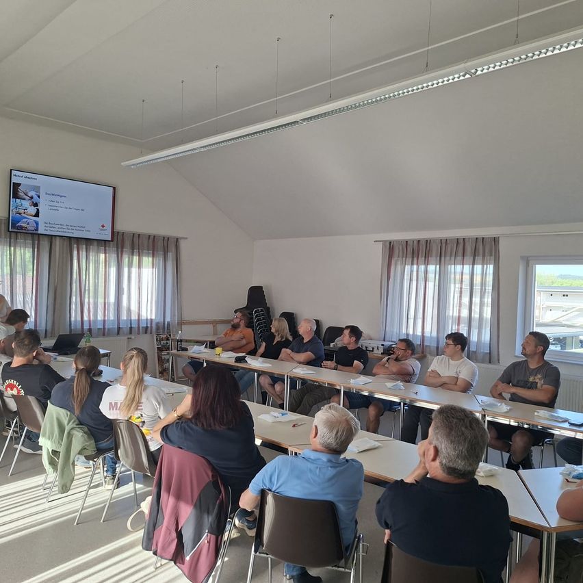 Eine Gruppe von Menschen sitzt in einem Klassenzimmer. Einige tragen Brillen, und eine Projektionsleinwand befindet sich an der Wand. Es gibt mehrere Tische und Stühle, und ein Heizkörper ist an der Wand. An der Seite befinden sich Fenster mit Vorhängen.