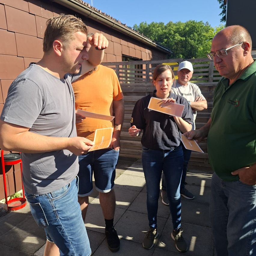 Fünf Personen stehen in einem Außenbereich und halten Papiere in der Hand. Der Mann auf der linken Seite hält ein Papier in der rechten Hand. Eine andere Person auf der rechten Seite hält ein Papier in beiden Händen. Sie alle schauen auf das Papier. Hinter ihnen befindet sich ein Holzzaun.