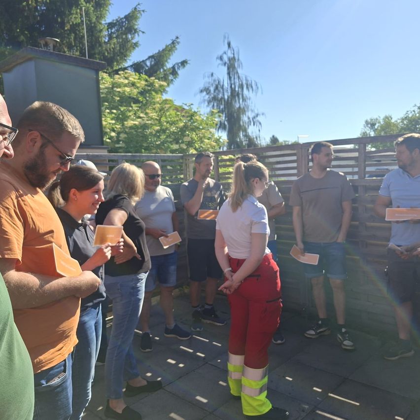 Eine Gruppe von Menschen steht in einer Reihe und hält orangefarbene Papiere in der Hand, möglicherweise unterhalten sie sich. Eine Frau in roten Hosen schaut auf etwas am Boden. Hinter ihnen steht ein Holzzaun mit Bäumen und einem klaren blauen Himmel.