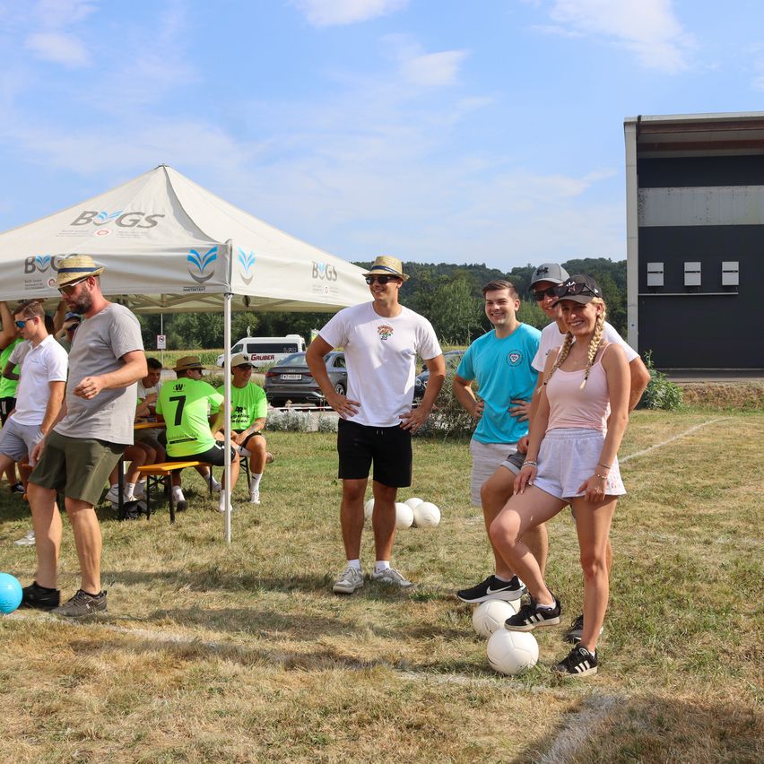 Eine Gruppe von Menschen spielt ein Spiel mit Fußballbällen auf einem Rasenfeld unter einem Zelt. Eine Person steht auf den Bällen.