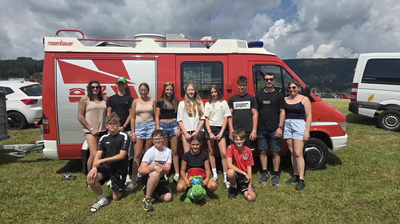 A group of people, including children, are posing for a photo in front of a red fire truck. The fire truck is parked on a grassy field with a mountain range in the background.