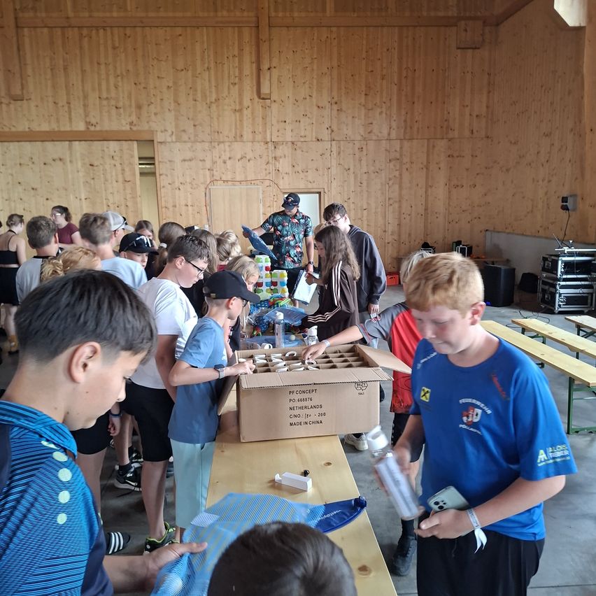 A group of young people are gathered around a table with a box of items, possibly preparing for an event. Some are wearing hats and glasses, and others are holding items. The room has wooden walls and a wooden floor.