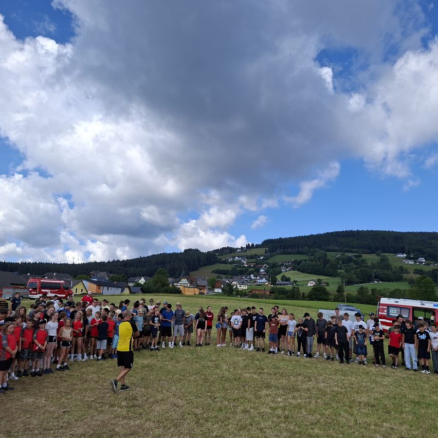 A large group of people, possibly students, are gathered in a field under a partly cloudy sky. A man in a yellow shirt is walking in front of them. In the background, there are houses and a mountain range.