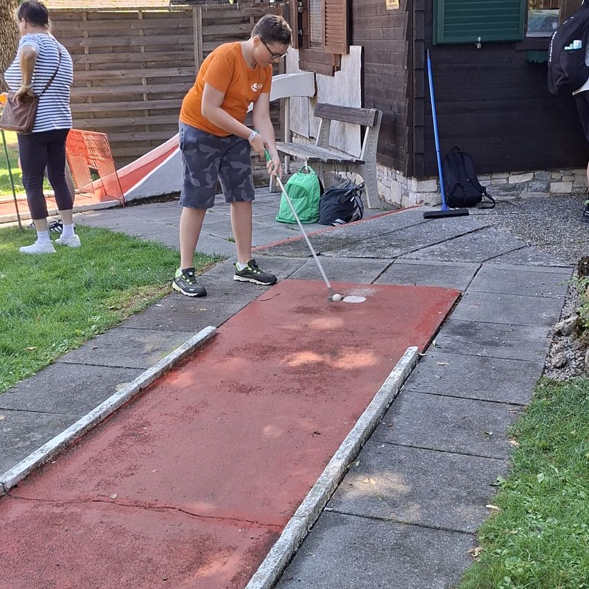 A young man in an orange shirt is playing mini golf on a red course outside.