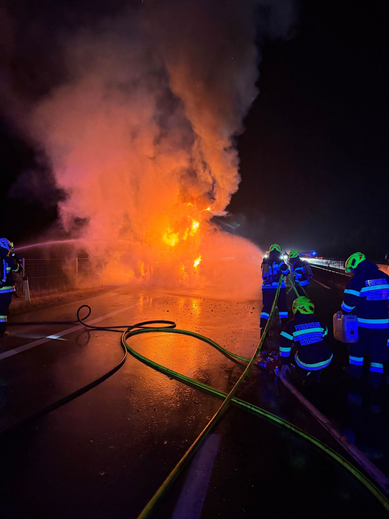 Feuerwehrleute in Schutzkleidung bekämpfen ein großes Feuer auf einer Straße bei Nacht, einer spritzt Wasser und andere arbeiten mit Schläuchen.