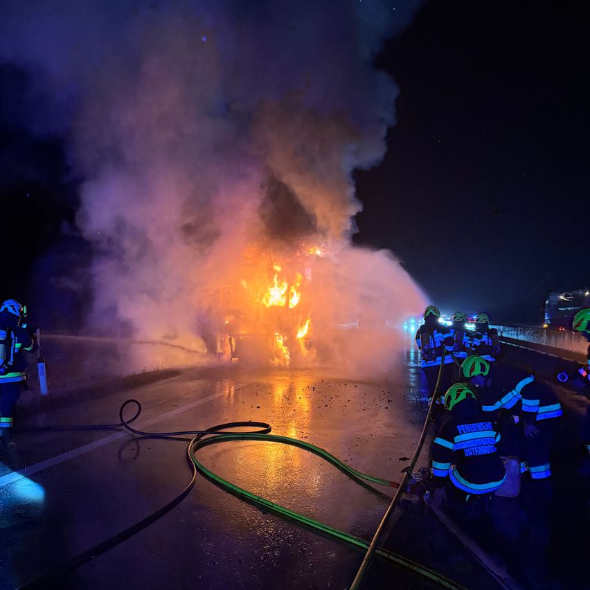 Feuerwehrleute löschen ein brennendes Fahrzeug auf einer Straße bei Nacht, mit Rauch und Wasser, das aus der Szene spritzt.