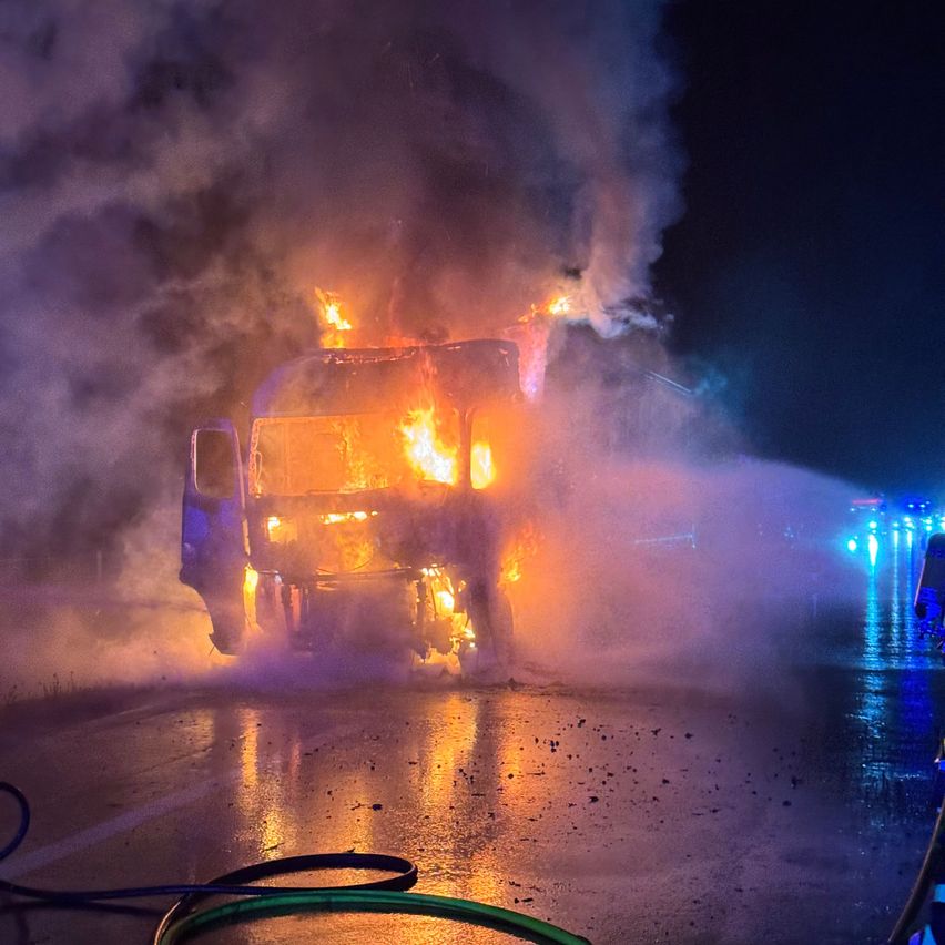 Ein brennender Lkw auf einer nassen Straße bei Nacht, mit Feuerwehrleuten, die Wasser sprühen, und blinkenden Notfallleuchten.
