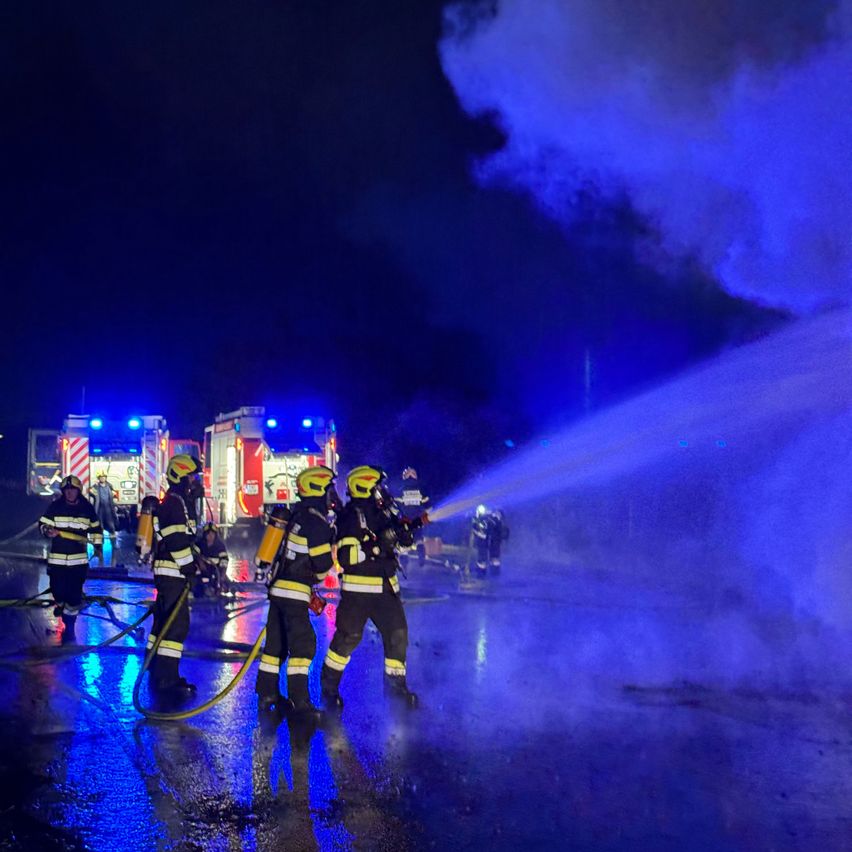 Feuerwehrleute in Schutzkleidung sprühen Wasser auf ein Feuer bei Nacht, mit Feuerwehrwagen und blinkenden Lichtern im Hintergrund.