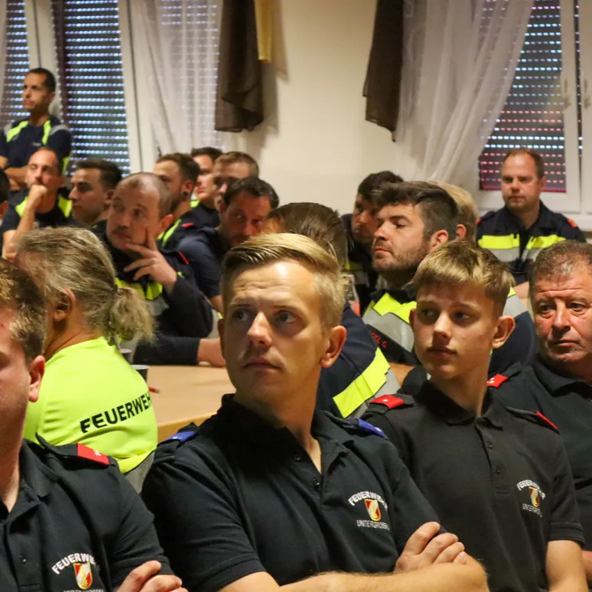 Eine Gruppe von Feuerwehrleuten in Uniform sitzt in einem Raum, einige schauen nach vorne, andere nach unten. Der Raum hat weiße Wände und Fenster mit Jalousien.