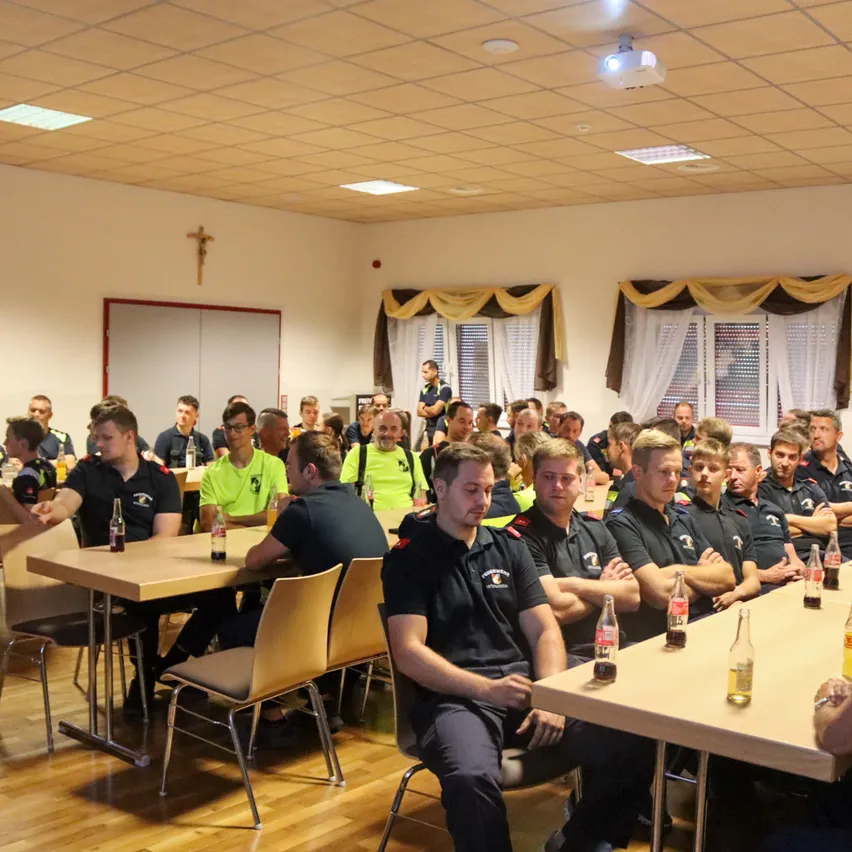 Eine Gruppe von Feuerwehrleuten in Uniform sitzt an Tischen in einem Raum mit Holzböden und weißen Wänden. Flaschen und Dosen sind auf den Tischen.