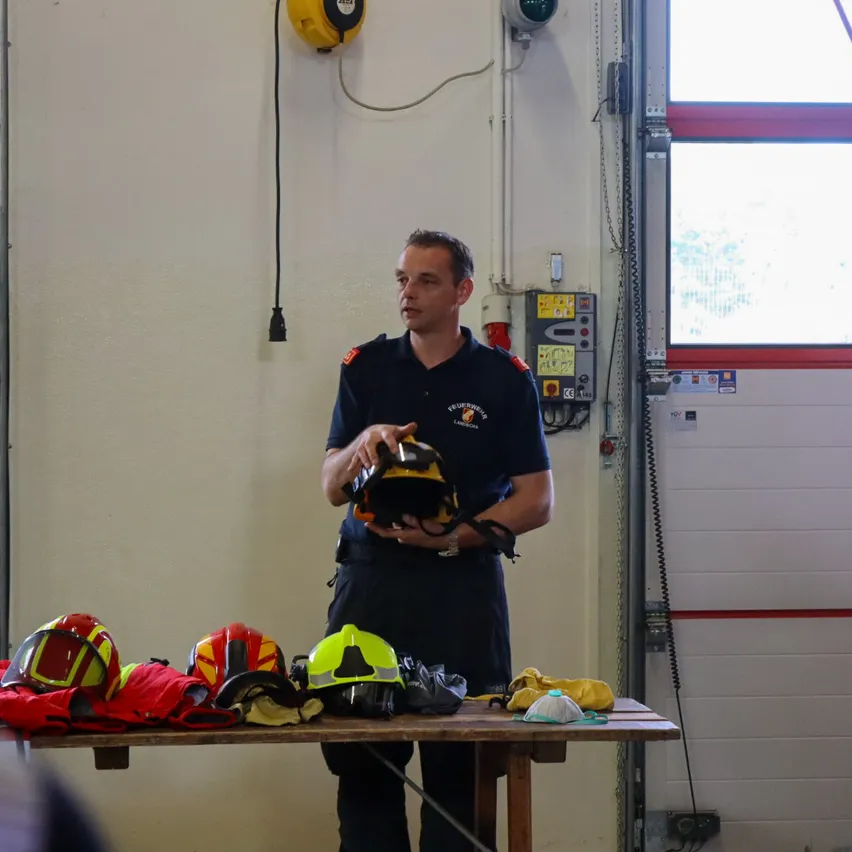 Ein Feuerwehrmann steht vor einem Tisch mit verschiedenen Feuerwehrhelmen und Ausrüstung und hält einen Helm in den Händen.