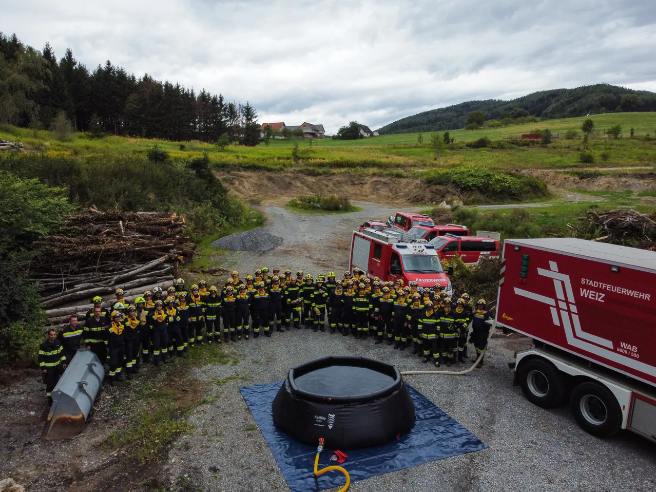 Eine Gruppe Feuerwehrleute in Uniform steht im Kreis um einen großen aufblasbaren Pool, mit Feuerwehrfahrzeugen und Ausrüstung im Hintergrund.