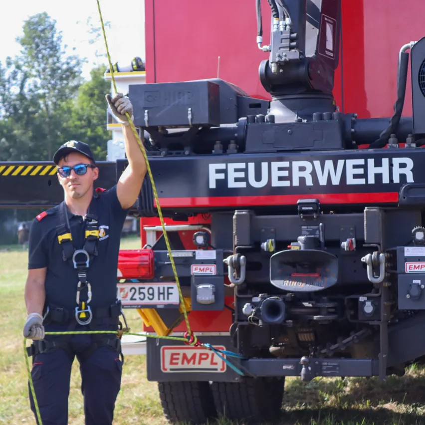 Ein Feuerwehrmann in voller Ausrüstung hält ein Seil, das an einem großen roten Feuerwehrfahrzeug mit der Aufschrift 'Feuerwehr' befestigt ist.