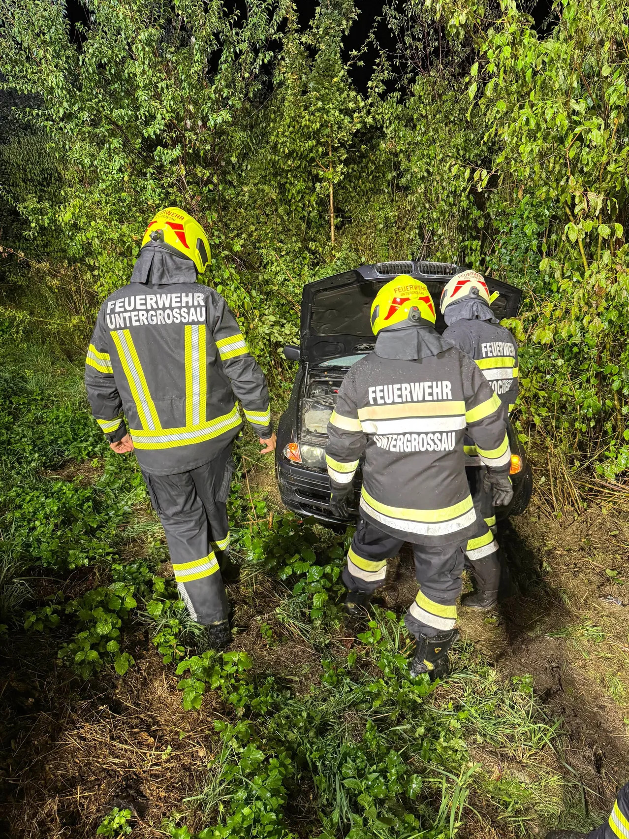Drei Feuerwehrleute in voller Ausrüstung arbeiten an einem Auto mit offenem Motorhaube in einem Waldgebiet.
