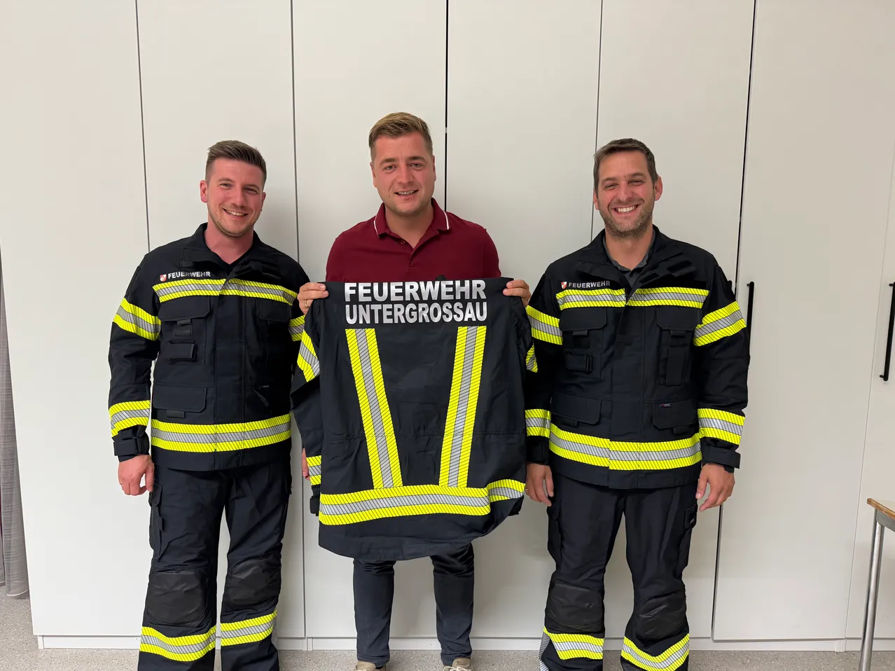Drei Feuerwehrleute in Uniform stehen zusammen, einer hält eine Jacke mit den Worten 'Feuerwehr Untergrossau'.