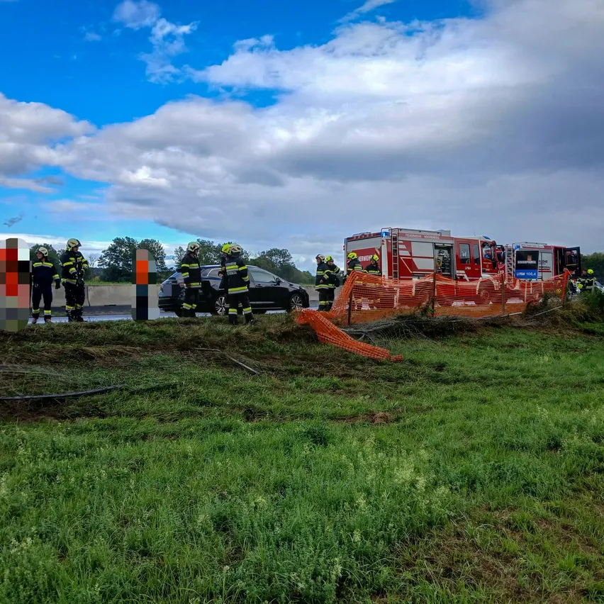 Rettungskräfte in Schutzkleidung stehen neben einer beschädigten Barriere und einem schwarzen Auto auf einem Grasfeld. Ein roter Feuerwehrwagen ist in der Nähe geparkt.