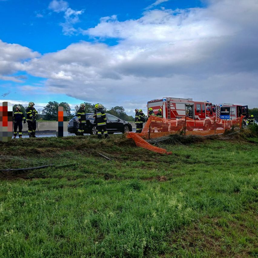 Rettungskräfte in Schutzkleidung stehen neben einer beschädigten Barriere und einem schwarzen Auto auf einem Grasfeld. Ein roter Feuerwehrwagen ist in der Nähe geparkt.