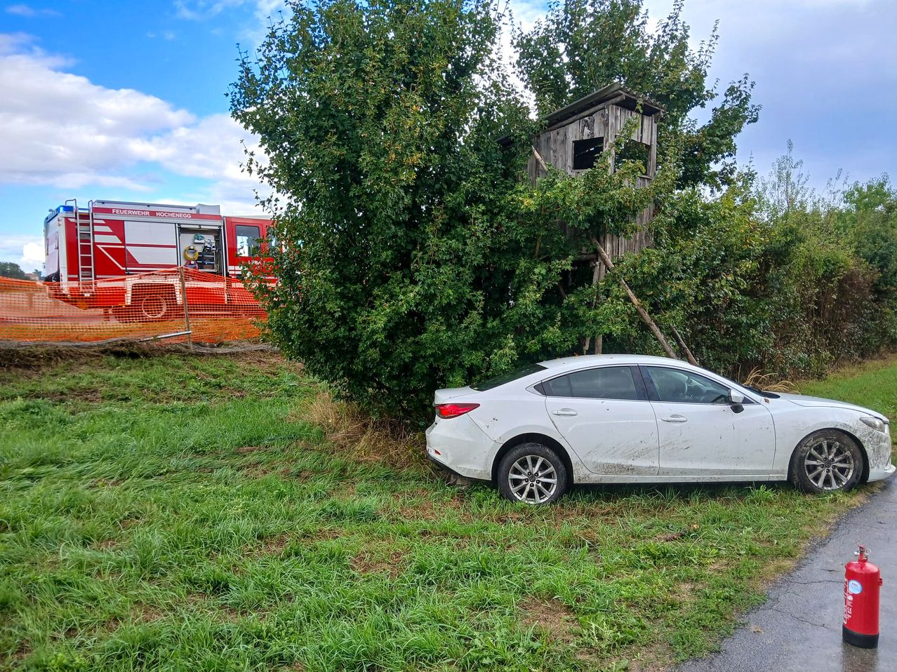 Ein weißes Auto ist in der Nähe eines Baumes geparkt, auf dem sich eine kleine Holzhütte befindet, mit einem Feuerwehrwagen im Hintergrund.