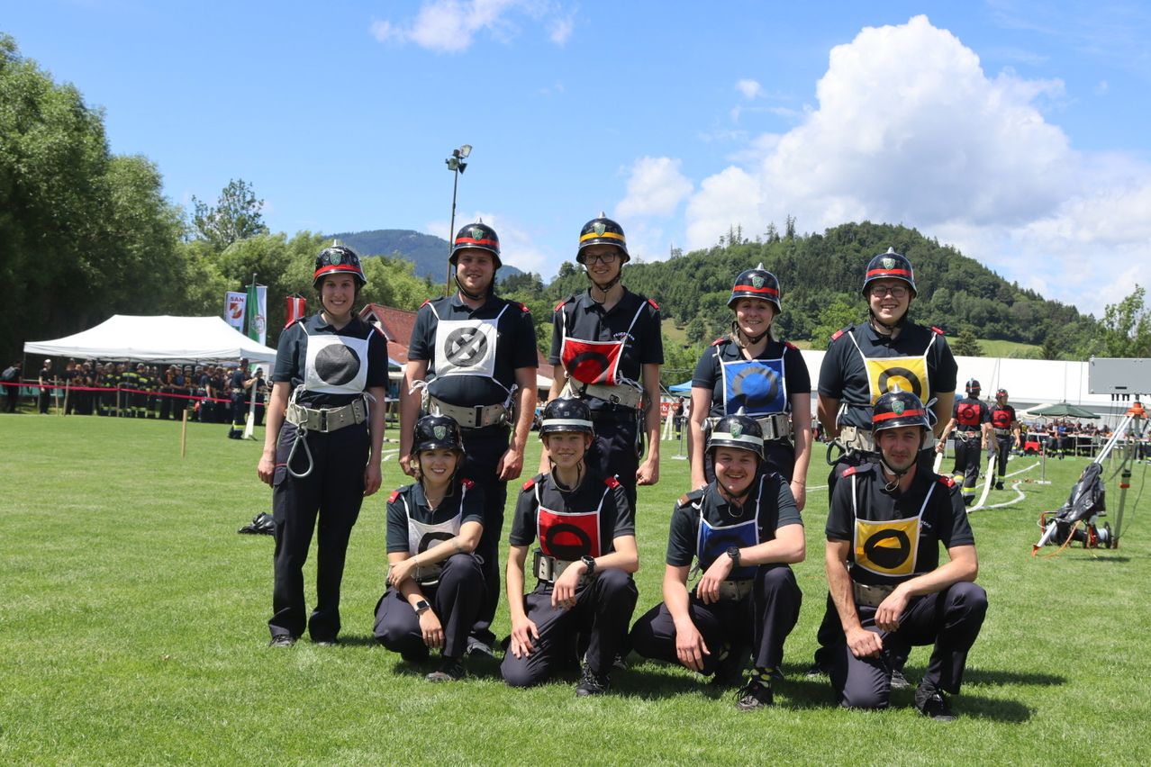Eine Gruppe von Personen, die Uniformen, Helme und Gürtel tragen, posiert für ein Foto auf einem Rasenfeld, mit Bäumen und Bergen im Hintergrund.