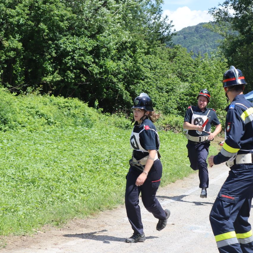 Drei Feuerwehrleute in Uniformen und Helmen rennen auf einem Pfad in einem Wald. Der vorne trägt die Nummer 8.