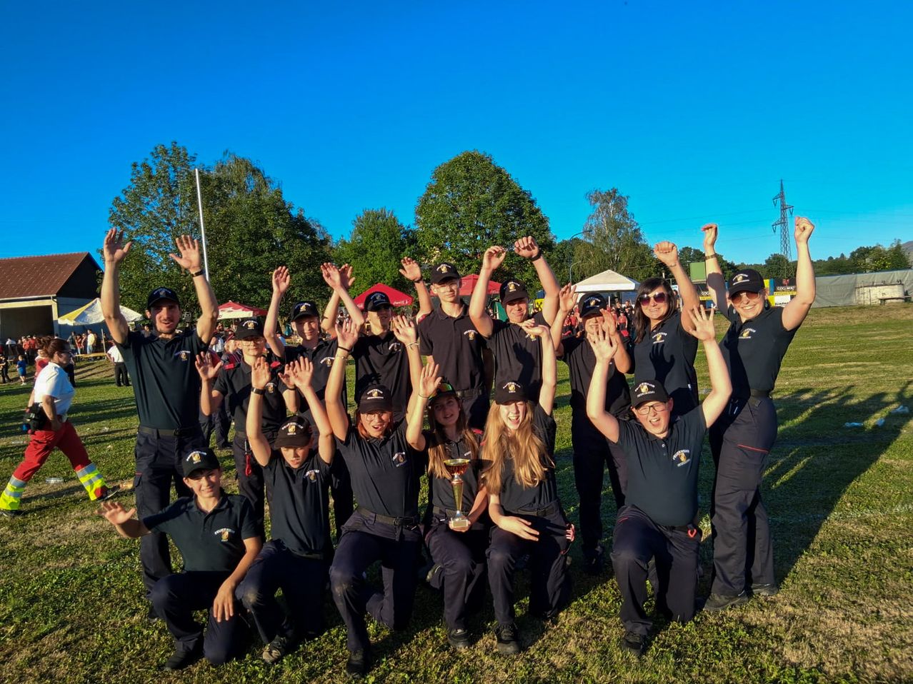 Eine Gruppe junger Erwachsener in Uniform posiert für ein Foto auf einem Feld mit einem Pokal. Sie haben ihre Hände erhoben und lächeln. Hinter ihnen sind Bäume und ein klarer blauer Himmel.