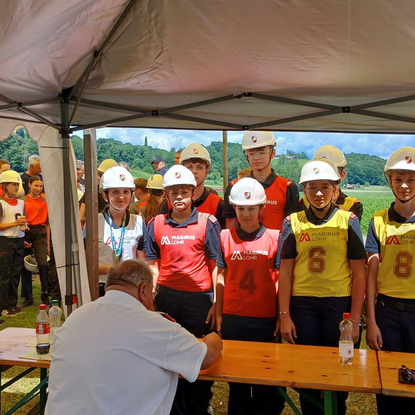 Eine Gruppe von Personen in Helmen und Uniformen steht unter einem Zelt. Sie nehmen wahrscheinlich an einer Schulveranstaltung teil. Ein Mann in einem weißen Hemd sitzt an einem Tisch und schreibt.