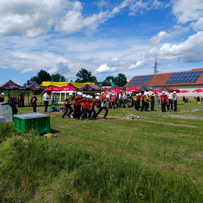 Eine Gruppe von Menschen in roten Uniformen und weißen Helmen steht auf einem Rasenfeld mit roten Zelten und Solarmodulen auf dem Dach im Hintergrund.