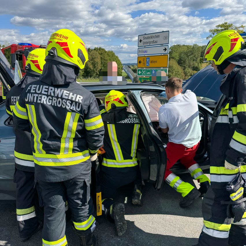 Feuerwehrleute in gelben und schwarzen Uniformen helfen einem Mann, der aus einem beschädigten Fahrzeug am Straßenrand aussteigt. Die Szene spielt sich unter einem teilweise bewölkten Himmel ab.
