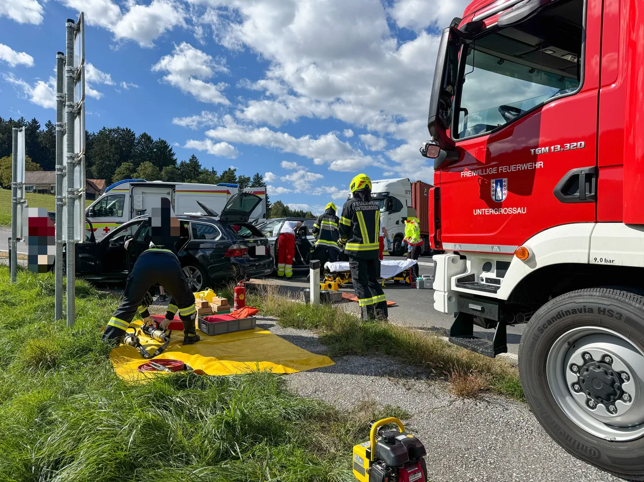 Feuerwehrleute kümmern sich um einen Autounfall an einem sonnigen Tag. Ein Feuerwehrmann benutzt ein Werkzeug auf dem Boden, während andere stehen. Ein Feuerwehrwagen und ein Krankenwagen sind in der Nähe geparkt.