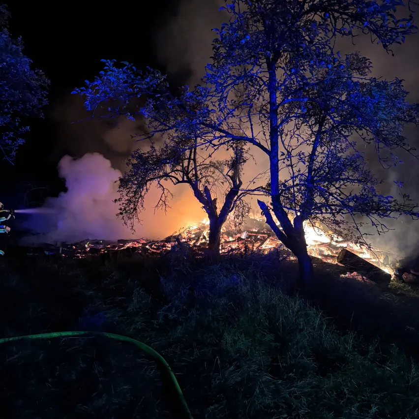 Eine nächtliche Szene mit einem Feuer in einem Feld, das von blauen Lichtern beleuchtet wird. Bäume umgeben das Feuer, und Rauch steigt in den dunklen Himmel auf.