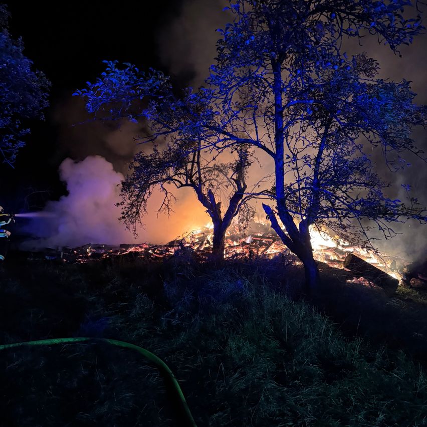 Eine nächtliche Szene mit einem Feuer in einem Feld, das von blauen Lichtern beleuchtet wird. Bäume umgeben das Feuer, und Rauch steigt in den dunklen Himmel auf.