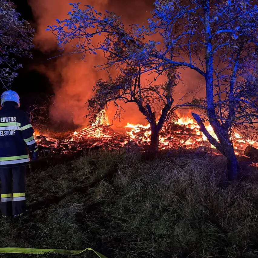 Ein Feuerwehrmann in Uniform steht neben einem brennenden Holzhaufen auf einem Feld bei Nacht.