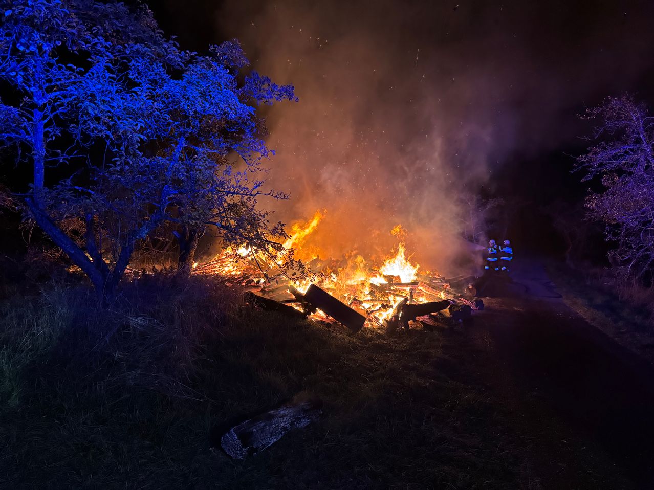 Ein großes Lagerfeuer brennt bei Nacht, umgeben von Bäumen und Gras, mit Notfallleuchten, die die Szene beleuchten.
